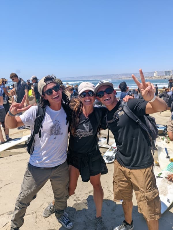 Three friends throwing peace signs on a crowded beach, surfboards on the sand behind them